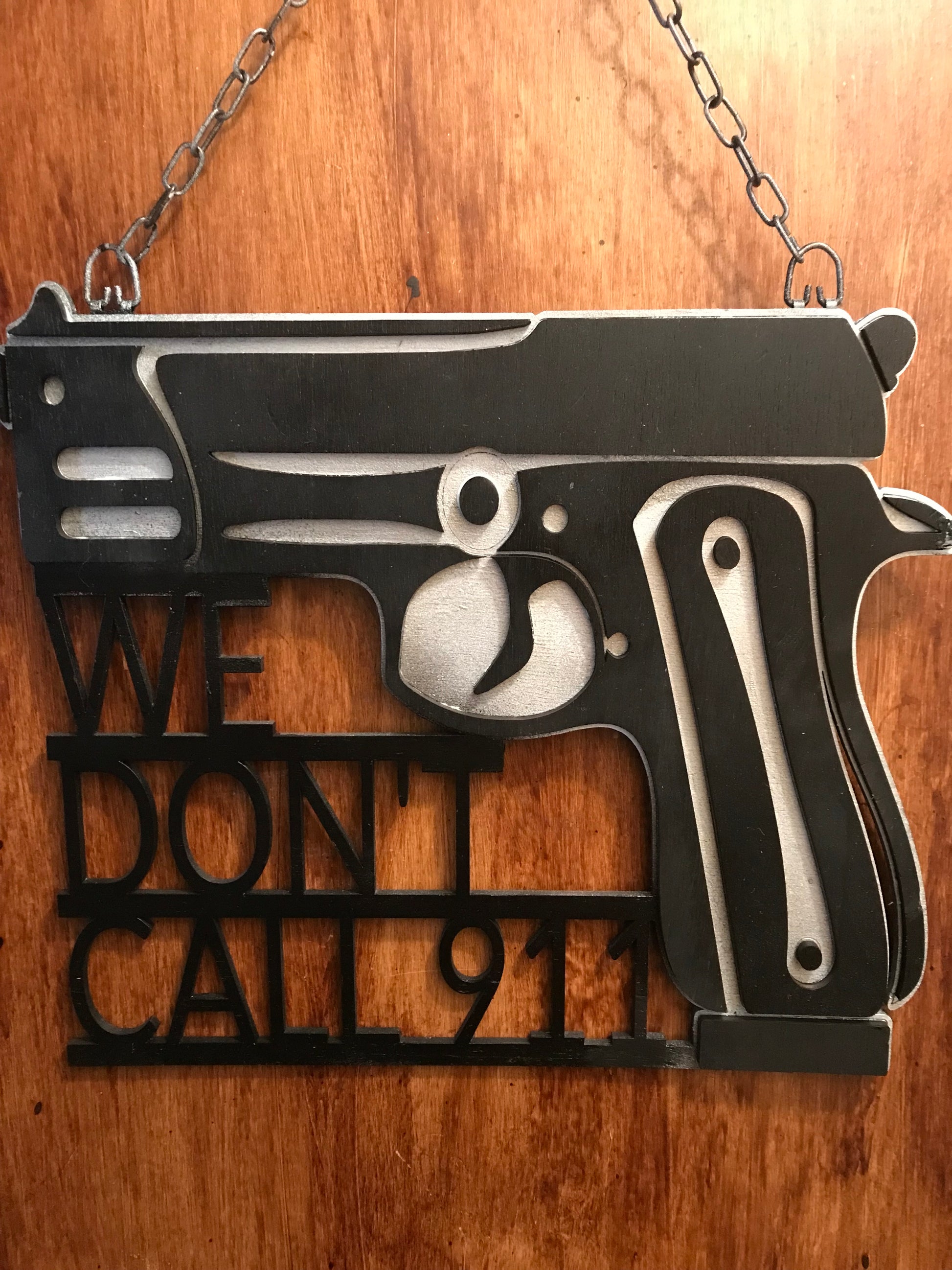 Picture of wooden gun door hanger with the words we don’t call 911. The background of the sign is silver with black details and lettering. The sign is hanging on a wooden door.